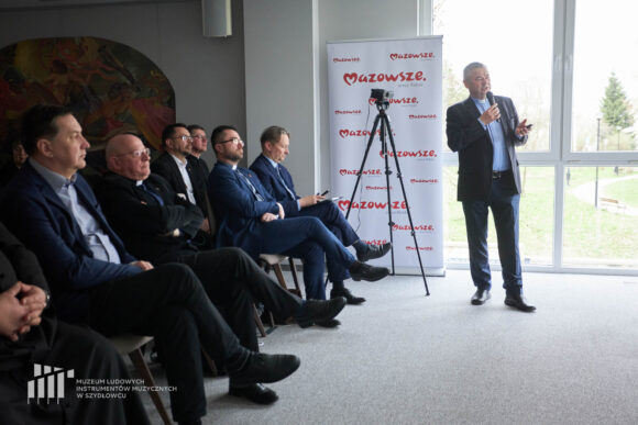 Pierwszy rząd słuchaczy, mężczyźni w ciemnych garniturach. Ksiądz zwórcony w kierunku prezentacji, stoi i przemawia przez mikrofon.