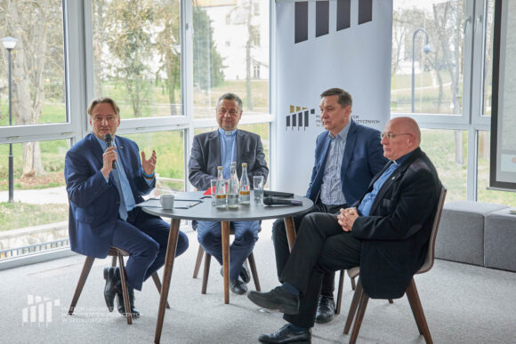 Zbliżenie na czterech mężczyzn siedzących przy stoliku, jeden z nich trzyma mikrofon i patrzy w kierunku aparatu.