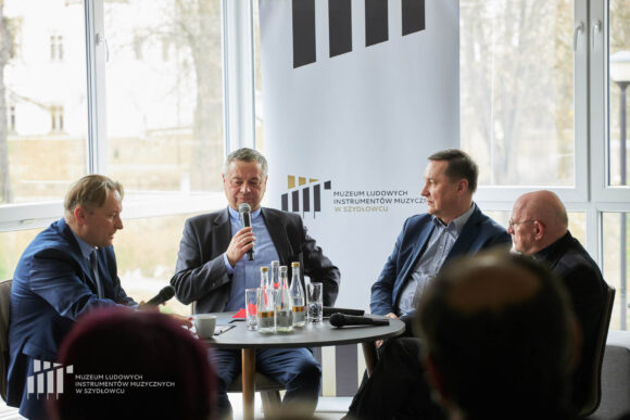 Zbliżenie na czterech mężczyzn siedzących przy stoliku, jeden z nich trzyma mikrofon.