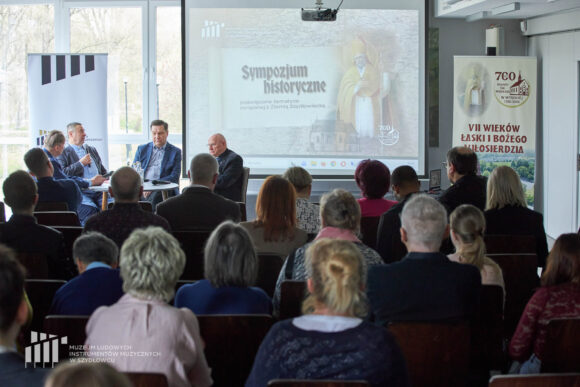 Czterech mężczyzn siedzących przy stoliku podczas debaty, przed nimi publiczność, za nimi na ekranie prezentacja multimedialna oraz dwa rollupy.