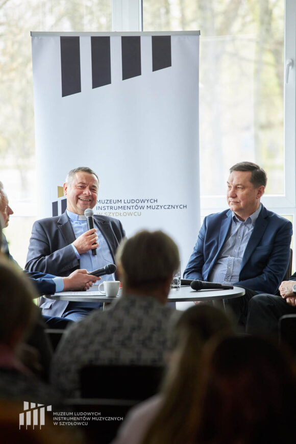 Dwóch mężczyzn przy stoliku. Jeden z nich uśmiecha się i mówi przez mikrofon. Za nimi rollup Muzeum w Szydłowcu oraz okna z dziennym światłem.