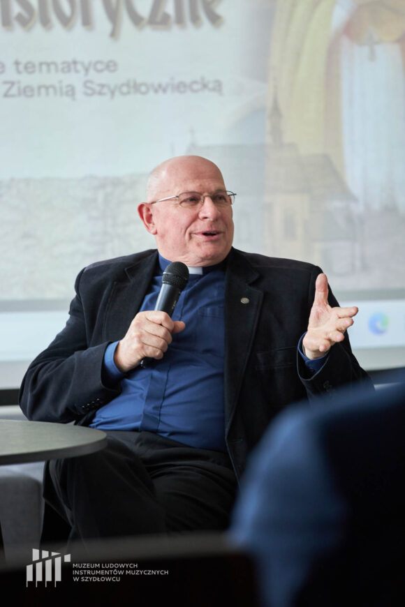 Ksiądz w granatowej koszuli z koloratką w czarnej marynarce siedzi i przemawia przez mikrofon.