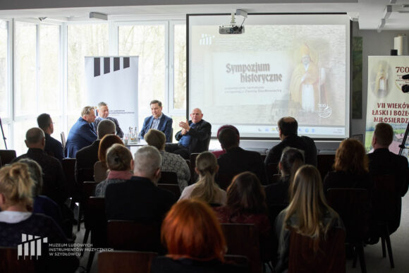 Czterech mężczyzn siedzących przy stoliku podczas debaty, przed nimi publiczność, za nimi na ekranie prezentacja multimedialna oraz dwa rollupy.