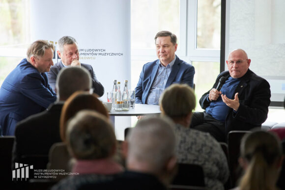 Zbliżenie na czterech mężczyzn siedzących przy stoliku, jeden z nich trzyma mikrofon i patrzy w kierunku publiczności przed nimi.
