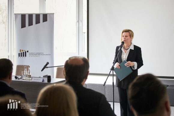 Zbliżenie na kilka osób z publiczności oraz dyrektor muzeum przemawiającą przez stojący przed nią mikrofon. Trzyma w ręku papierową teczkę.