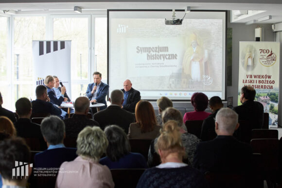 Czterech mężczyzn siedzących przy stoliku podczas debaty, przed nimi publiczność, za nimi na ekranie prezentacja multimedialna oraz dwa rollupy.