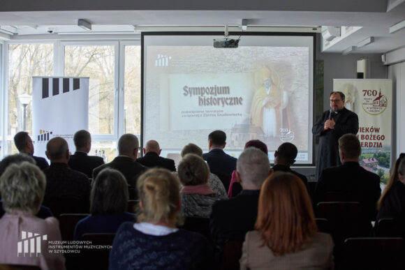 Po prawej stronie ksiądz przemawiający przez mikrofon, na środku ekran z grafiką sympozjum historycznego, po lewej okna z dziennym światłem i rollup Muzeum w Szydłowcu. Na pierwszym planie pierwsze rzędy publiczności.