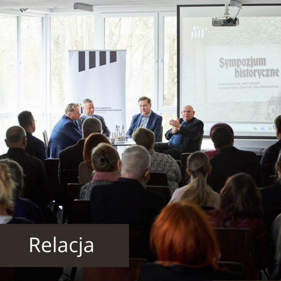 Czterech mężczyzn siedzących przy stoliku podczas debaty, przed nimi publiczność.