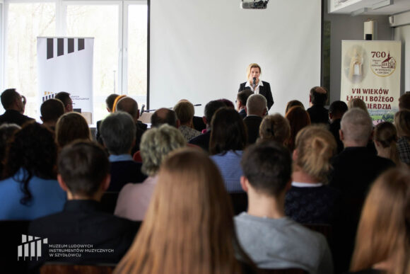 Dyrektor muzeum przemawiająca przez stojący przed nią mikrofon. Bliżej sylwetki siedzącej na krzesłach publiczności w różnym wieku, od młodzież po seniorów.
