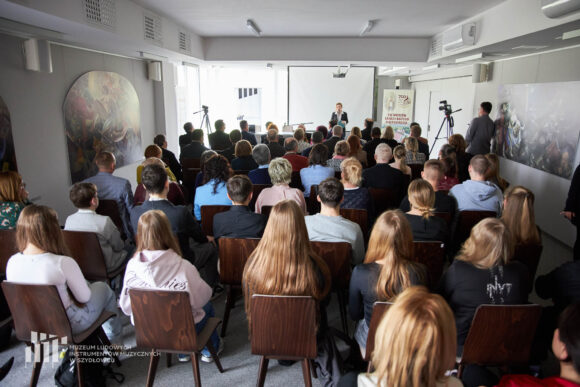 Widok z końca sali konferencyjnej. W oddali dyrektor muzeum przemawiająca przez stojący mikrofon. Bliżej siedząca na krzesłach publiczność w różnym wieku, od młodzież po seniorów.