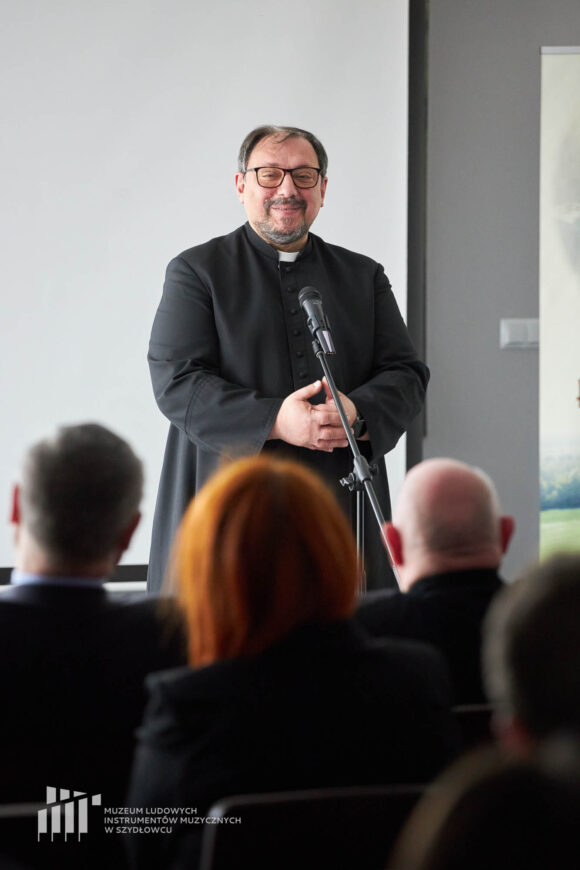 Zbliżenie na sylwetkę uśmiechniętego księdza w sutannie, ma złożone dłonie. Stoi przed mikrofonem. Przed nim rozmyte sylwetki siedzącej publiczności.