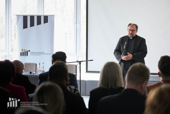 Zbliżenie na księdza przemawiającego przy stojącym przed nim mikrofonie do siedzącej publiczności. Po lewej biały rollup Muzeum w Szydłowcu. Za księdzem biały ekran.