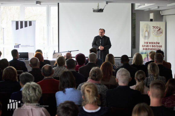 Na pierwszym planie publiczność sympozjum. Na scenie ksiądz w czarnej sutannie, stoi i przemawia przed mikrofonem. Za nim biały ekran i dwa rollupy po bokach.