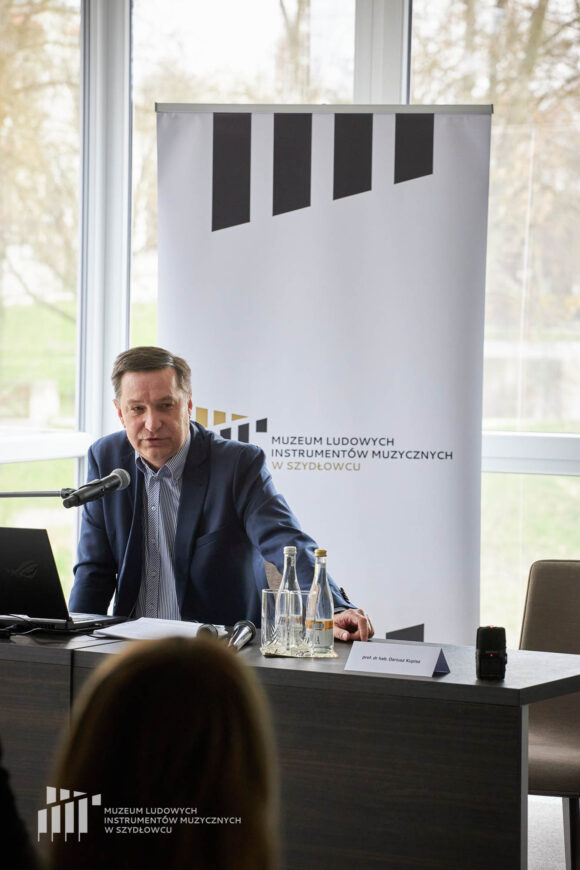 Prelegent siedzi przy stoliku przy mikrofonie przed publicznością. Na stoliku stoi woda w butelkach i szklanki, laptop, mokrofony i materiały. Za nim rollup Muzeum w Szydłowcu i okna ze światłem dziennym.