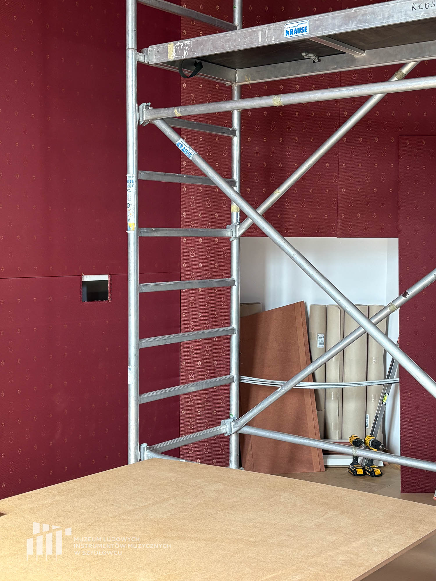 Prace budowlane przygotowujące do otwarcia nowej wystawy. Widać cześć rusztowania. Ściany zostały obite tkaniną w kolorze bordo.
