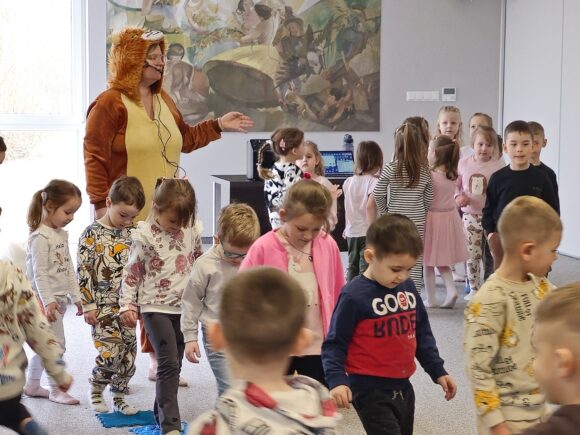 Animatorka w żółto brązowym stroju lwa z mikrofonem przy twarzy. Obok gromada dzieci biegających gęsiego po matach sensorycznych.