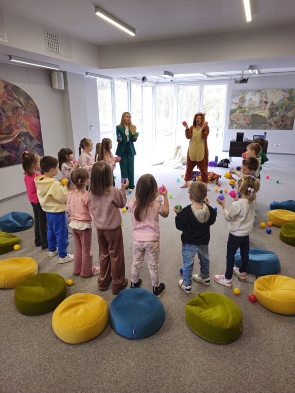 Animatorka w żółto brązowym stroju lwa z mikrofonem przy twarzy pokazuje zabawę z piłeczkami. Obok gromada dzieci ustawionych w kole, każdy na nich trzyma dwie piłki.