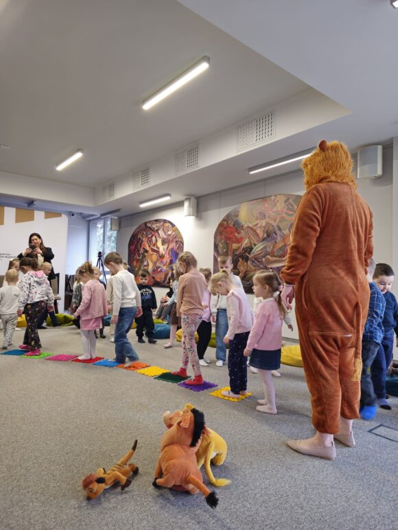 Animatorka w brązowym stroju lwa. Obok gromada dzieci biegających gęsiego po matach sensorycznych. w oddali kobieta robi zajęcie. Na podłodze pluszowe zabawki bohaterów.