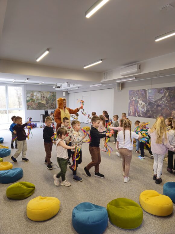 Gromada dzieci biegających ze wstążkami. Przed nimi kolorowe poduchy. Między nimi aktorka.