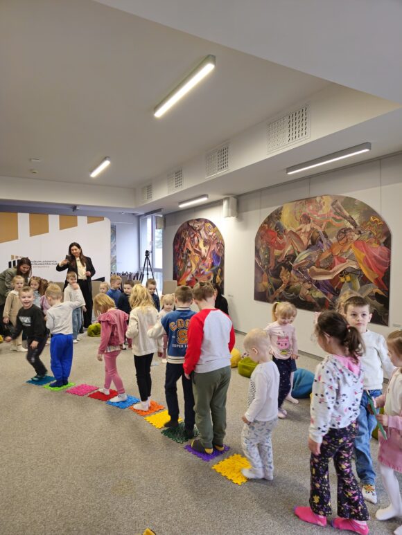 Dzieci biegają gęsiego po matach sensorycznych. W oddali nauczycielka robi im zdjęcia.