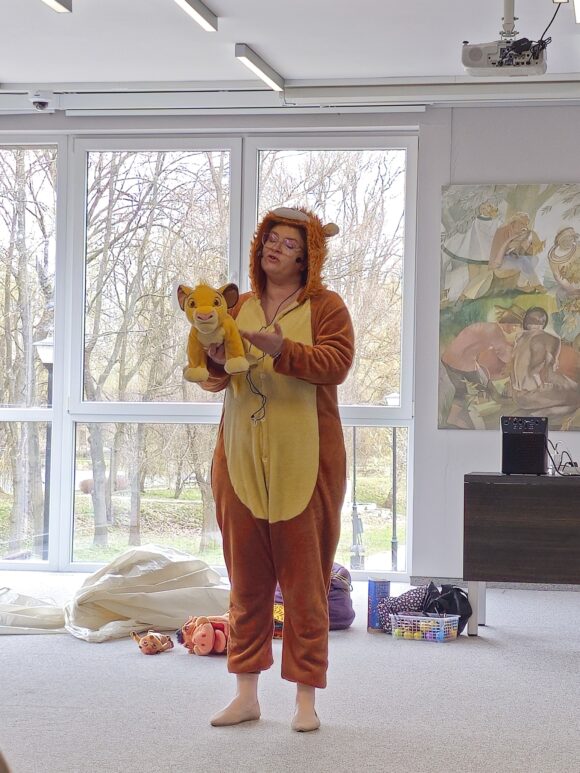 Aktorka w okularach w żółto brązowym stroju lwa z mikrofonem przy twarzy trzyma pluszowego misia, o którym opowiada historię. Za nią inne zabawki i akcesoria do spektaklu.