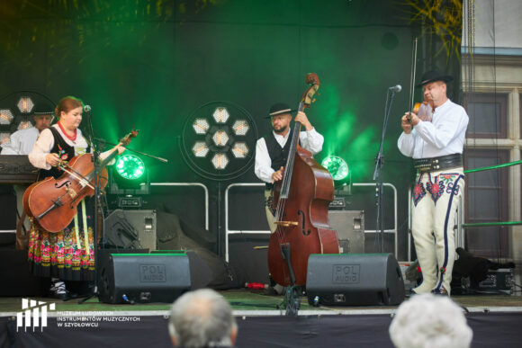Troje muzyków w strojach góralskich gra na różnych instrumentach.