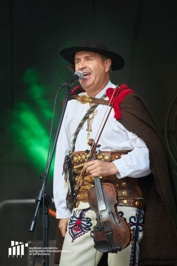 Zbliżenie na górala śpiewającego do mikrofonu. W lewej ręce trzyma skrzypce.