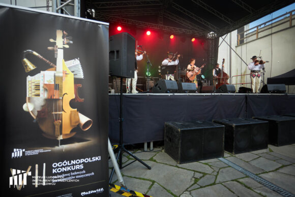 Po lewej stronie plakat VII Ogólnopolskiego Konkursu na Budowę Ludowych Instrumentów Muzycznych. Dalej po prawo, w pewnym oddaleniu muzycy na scenie.