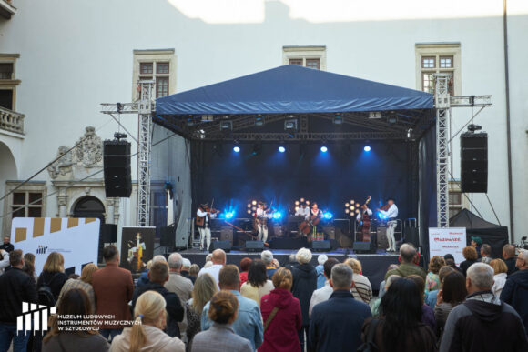 W oddali na wprost scena z grającymi na niej muzykami. Na pierwszym planie plecy ludzi oglądających koncert.