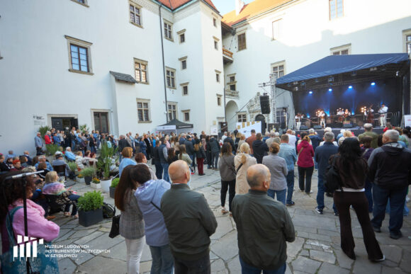 W tle białe muzy zamku. Po prawo scena z granatowym dachem. Na pierwszym planie ludzie stojący tyłem do patrzącego na zdjęcie.