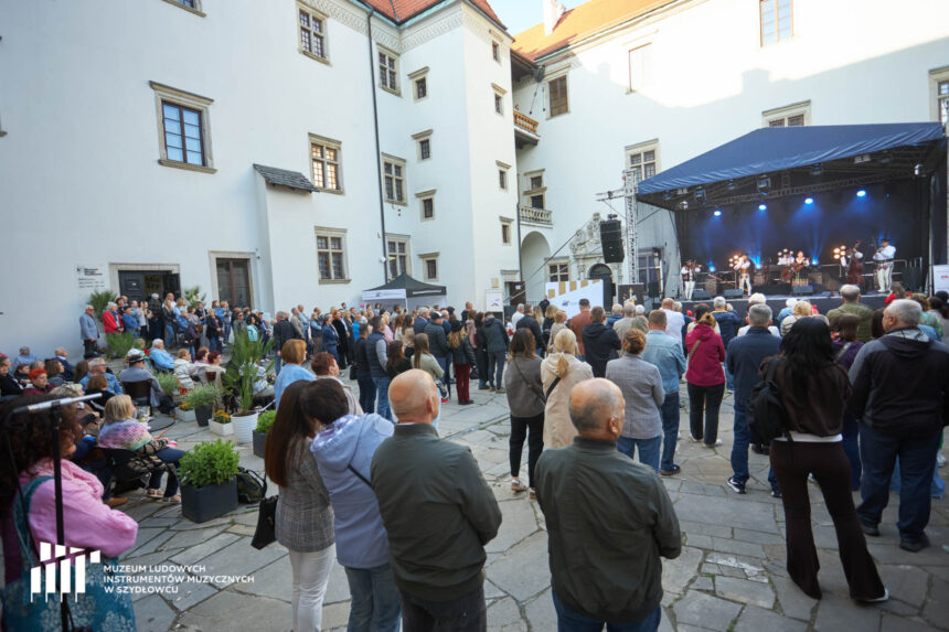 W tle białe muzy zamku. Po prawo scena z granatowym dachem. Na pierwszym planie ludzie stojący tyłem do patrzącego na zdjęcie.