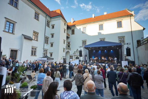 Dziedziniec zamku. Od lewej strony i na wprost widoczny zamek - białe otynkowane mury, okna w kamiennych oprawach, pomarańczowy, spadzisty dach. W dolnej części ludzi stojących na dziedzińcu, patrzą na scenę, na której gra kapela góralska. nad sceną granatowy dach.