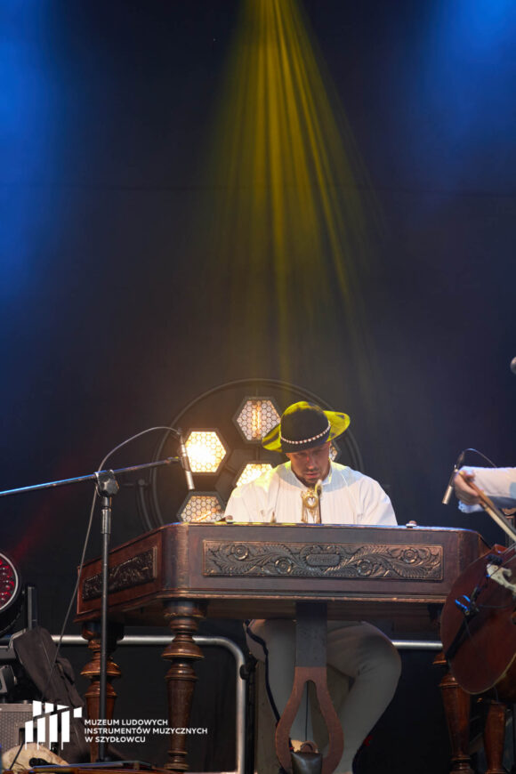 W dolnej części zdjęcia na pierwszym planie drewniany instrument muzyczny - przypominający kształtem pianino, wsparty na czterech nogach. Pozioma część zdobiona płaskorzeźbami. To cymbały węgierskie. Przy instrumencie siedzi mężczyzna w góralskim stroju. Tło ciemno-granatowe rozświetlone od góry żółtym światłem.