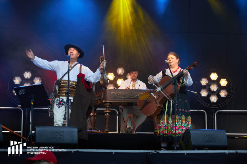 Trójka muzyków na scenie od góry oświetlonej żółtym światłem. Mężczyzna z lewej strony zdjęcia z rozłożonymi na boki, podniesionymi rękami, mówi do mikrofonu. Po prawej stronie kobieta grająca na basach. W tle mężczyzna siedzący za drewnianym zdobionym instrumentem - cymbałami.