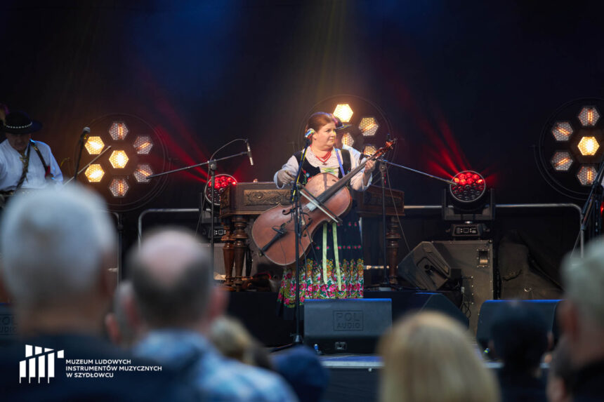 Na scenie stoi kobieta w góralskim stroju. Ma basy, które trzyma jak gitarę. W prawej ręce trzyma smyczek. Lewa ręka na gryfie. W tyłu światła sceniczne i ciemne tlo.