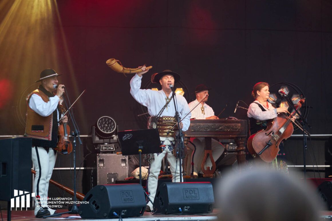 Koncert Trebunie-Tutki Na scenie widać trzy osoby w góralskich strojach. Mężczyzna w środku unosi prawą rękę, w której trzyma drewnianą trombitę.
