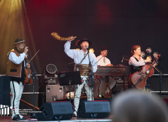 Koncert Trebunie-Tutki Na scenie widać trzy osoby w góralskich strojach. Mężczyzna w środku unosi prawą rękę, w której trzyma drewnianą trombitę.
