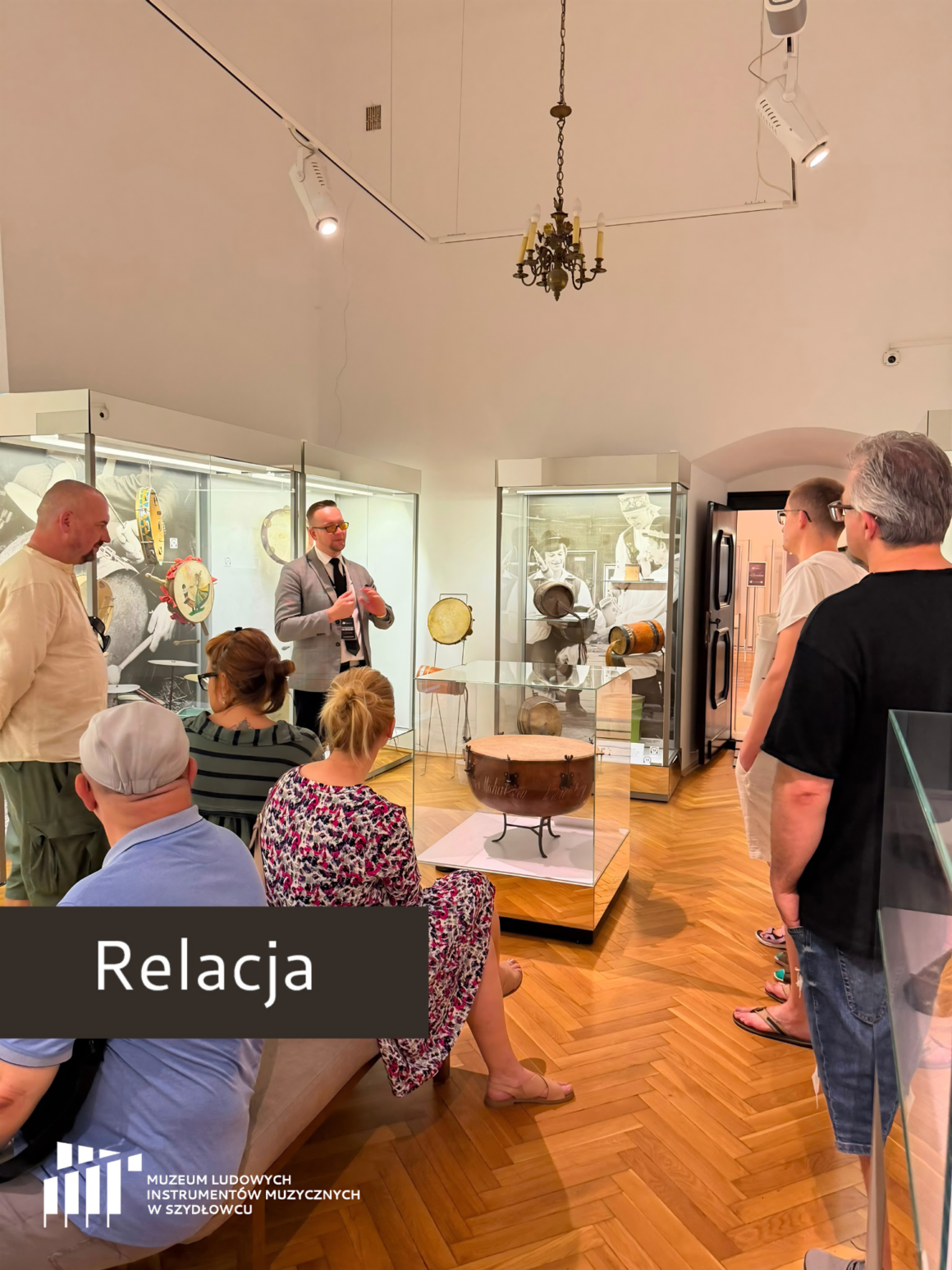 Muzeum Ludowych Instrumentów Muzycznych w Szydłowcu FolkRelaks - spacer z przewodnikiem. Na zdjęciu widać grupę ludzi stojących tyłem i przewodnika stojącego przodem do obrazu. Przewodnik jest ubrany w marynarkę, białą koszulę i krawat - ma okulary. W tle widac gablotę z instrumentami. Przeważa biały sufit.