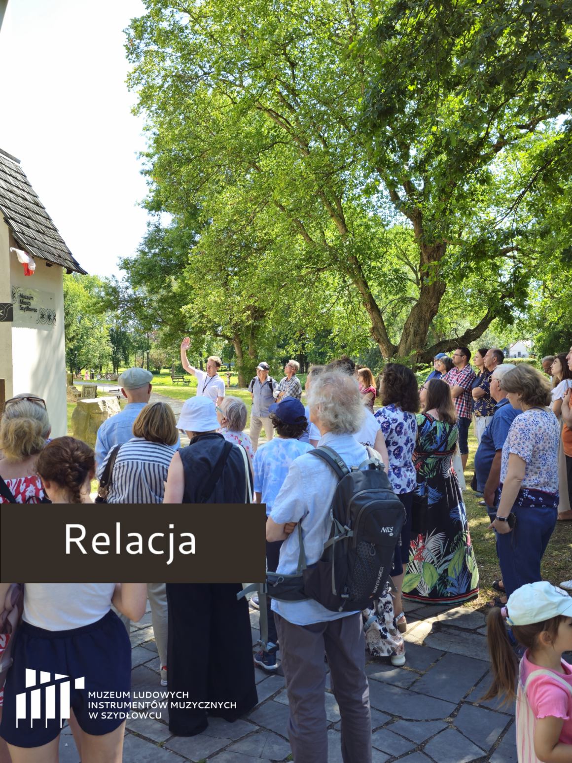 Projekt bez nazwy(3) Na pierwszym planie widać tłum ludzi stojących tyłem. W tle widać zielone drzewa i mury zamku.